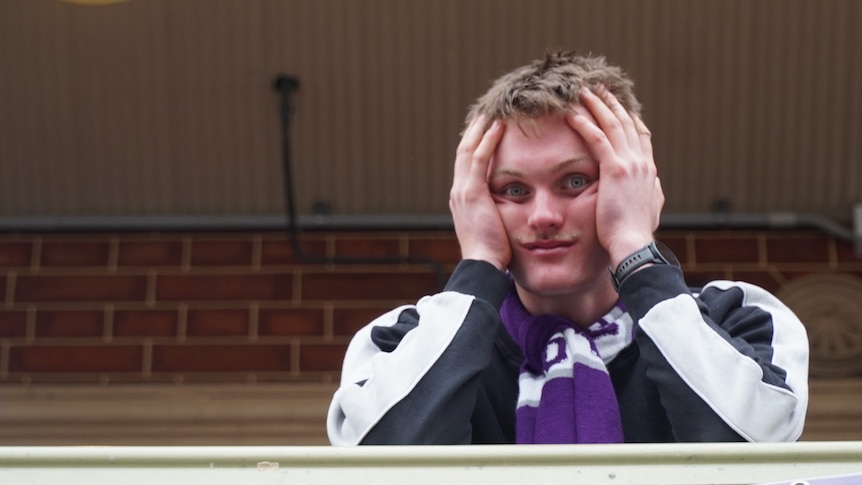 A man wearing a sports jacket and purple Fremantle scarf stares into a camera with his head in his hands