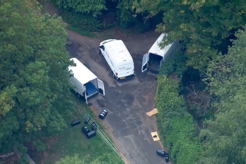 An aerial shot of three white vans, with two of their rear doors open, on a road near to multiple black barrels.