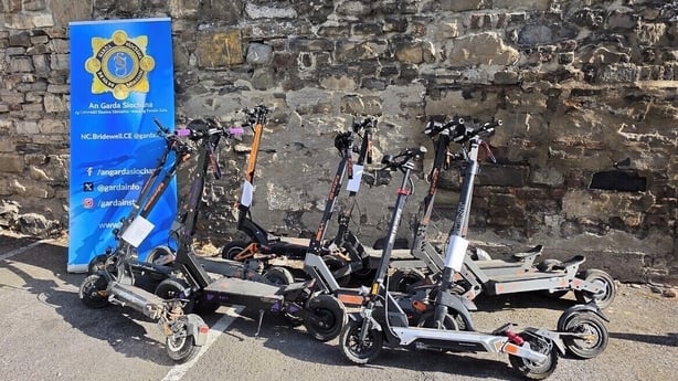 A number of e scooters are lined against a stone wall near a poster for the Gardaí